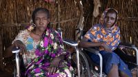 September 2024, Aboutengué, Chad. Kaltouma Ismail Abdullah (left) and her sister Hassaneih Ismail Abdullah (right) are both disabled. Thanks to the wheelchairs supplied by HI, they have regained their freedom of movement  Kaltouma and Hassaneih are side by side, each in a wheelchair. They are sheltered in a wooden house, looking at the lens. Katlouma, on the left, wears a dress with purple and white flowers, while Hassaneih, on the right, wears a blue scarf with orange flowers.