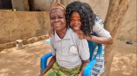 Likoulenan, a user of the African Network for Well-being and Resilience, and her mother. Savanes region, Togo. A young woman embraces an older woman sitting on a chair in a courtyard made of rammed earth. They both smile at the camera.