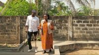 Souleyman Lawani arriving at the community rehabilitation centre and chatting with Prunelle Togni, HI's rehabilitation services officer in Benin. A man, wearing a prosthesis on his left leg, and a woman walk along a concrete path in a courtyard. The sun is shining brightly, and a wall and some vegetation can be seen behind them.