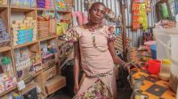 Poni Anet Wori, a refugee from South Sudan with a motor disability, standing in front of her shop in Kalobeyei camp, Kenya. A woman stands in a room made of sheet metal and wood. The walls are covered with shelves on which are arranged a variety of items: soap, candy, cookies, etc.