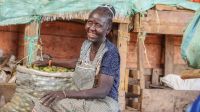 Esther Akuwam, a Kenyan woman living in Kakuma, manages a grocery shop. A woman sits laughing. Her right hand is resting on a large canvas bag containing fruit. Behind her are stalls of fruit and vegetables.
