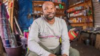 Ali Abdi Tifo, a Somali refugee living in the Kakuma refugee camp in Kenya, sitting in his shop. A man sits in a room whose walls and edges are covered with various objects: boxes, baskets, tarpaulins, ladles and so on. The man looks at the camera and smiles.