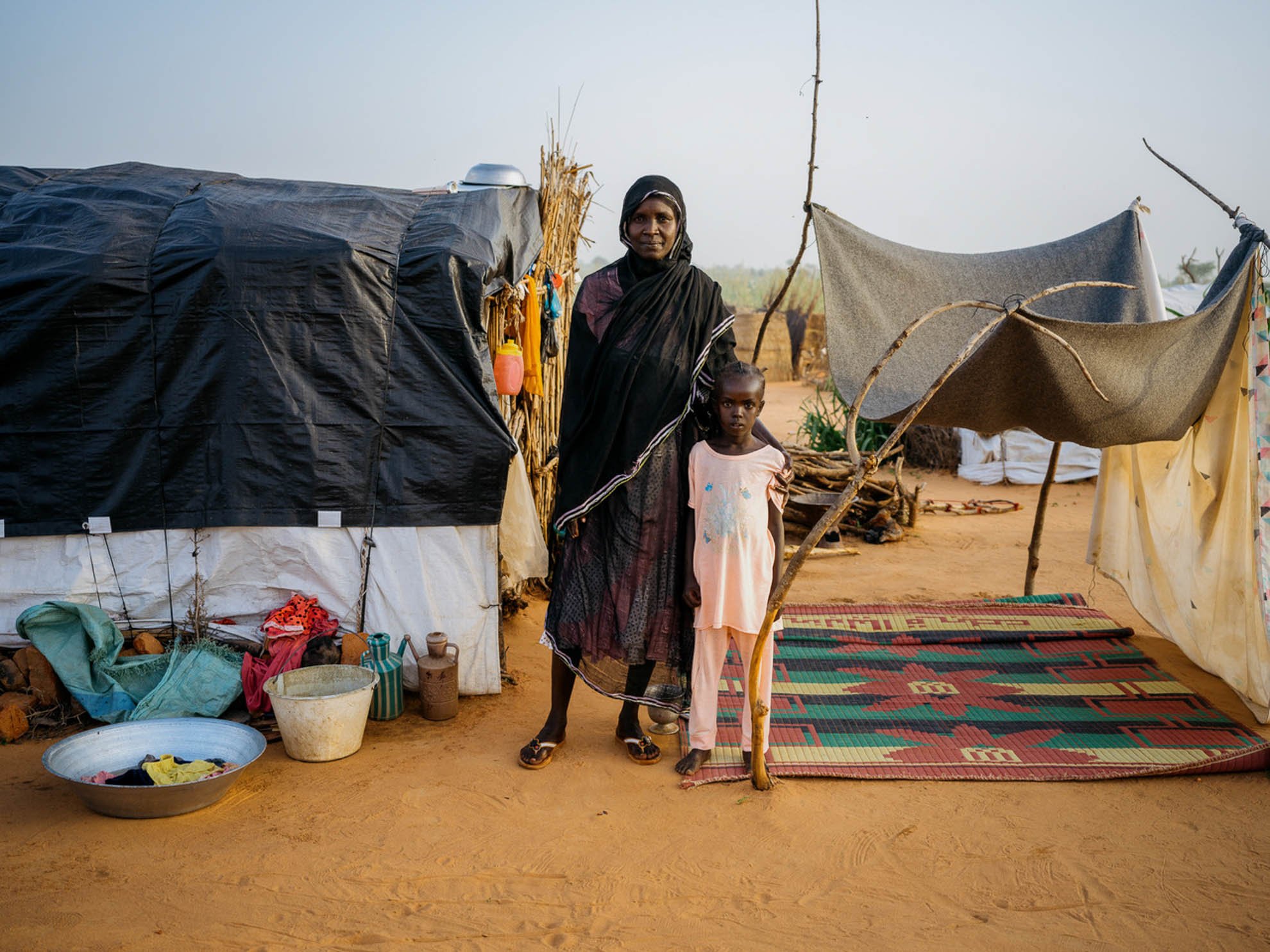 Nazik and Zahra are standing outside their shelter and looking at the camera