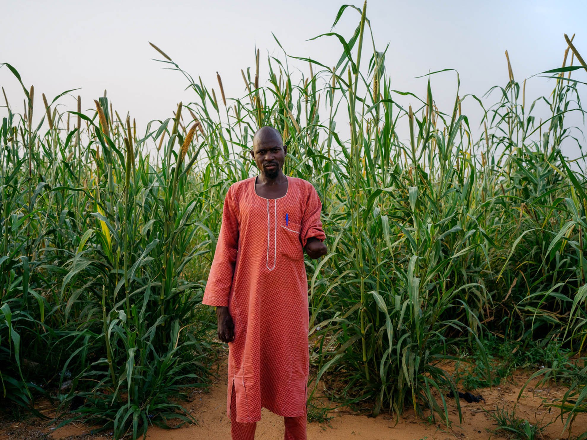 Mohamad is wearing a red outfit. His left arm has been amputated. He is standing up in front of high green plants