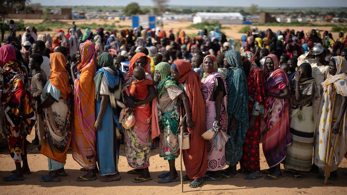 Des femmes se tiennent debout les unes derrière les autres, dans une file dont on distingue qu'elle continue loin derrière elles. Chacune tient à la main une bassine ou un récipient vide.