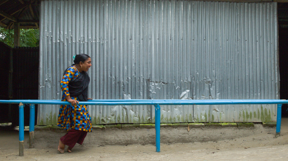 During a rehabilitation session, Anicha manages to walk and do her daily exercises with the help of these parallel bars, supplied by HI.The woman is standing on the frames looking at the camera with a big smile and wear a plaid skirt with different colors bleu orange yellow