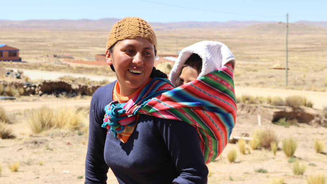 A smiling woman carries her son on her back using traditional aguayo cloth. In the background, the dry, dusty plains of the Altiplano.