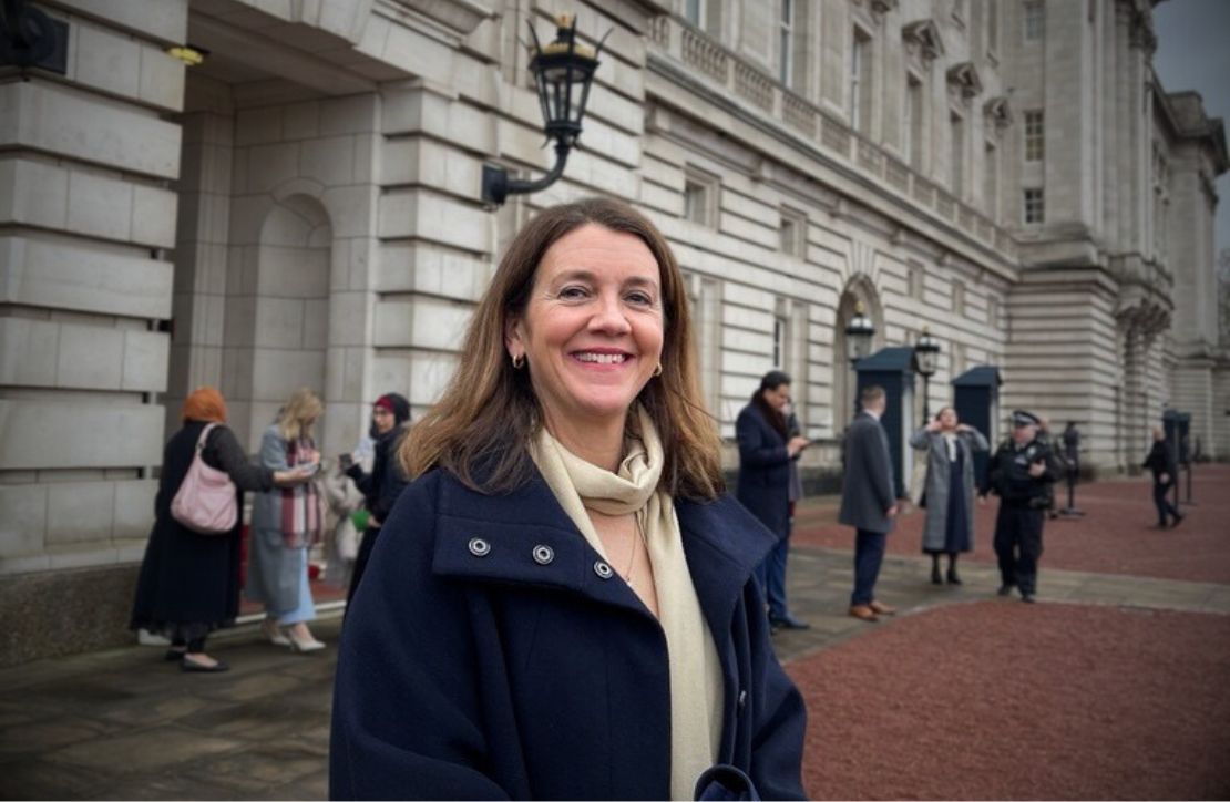 A portrait of Chloe Marshall standing outside Buckingham Palace. SHe is smiling and wearing a dark coat and a light scarf.